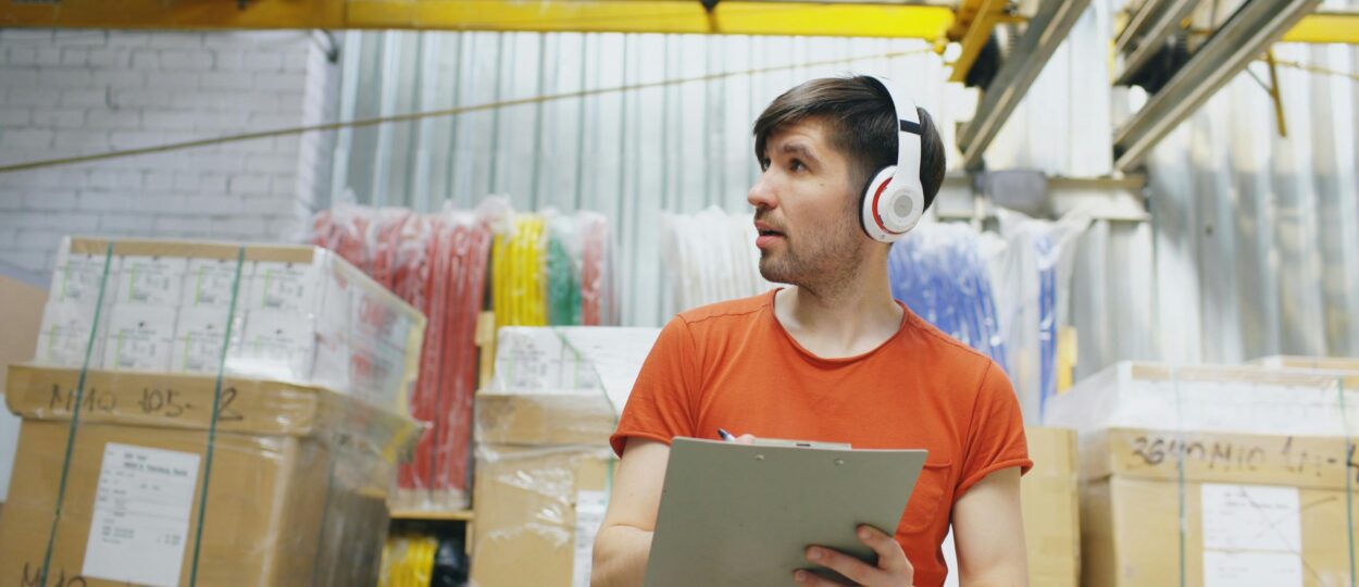 Young male warehouse worker with headphones checks inventory on clipboard in industrial setting.